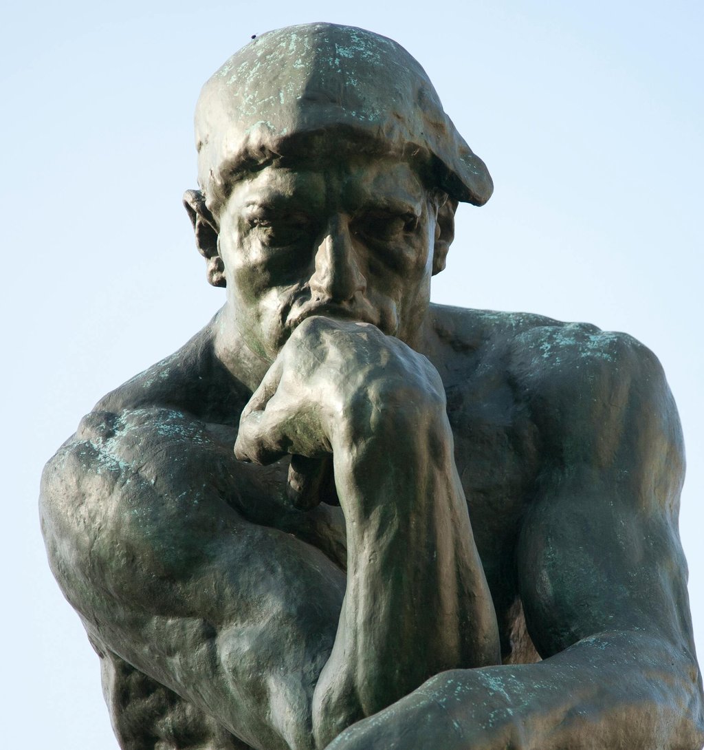 A close-up view of a bronze statue of Rodin's Thinker, depicting a muscular figure deep in thought, resting his chin on one hand. The statue exhibits detailed features and a contemplative expression, set against a clear blue sky.