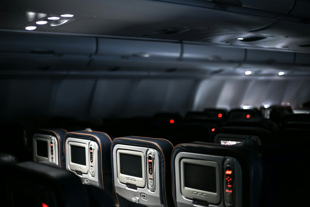 An empty airplane cabin features rows of seats with individual screens, dimly lit by overhead lights. The ambiance is tranquil, with soft blue and red lighting creating a serene atmosphere.