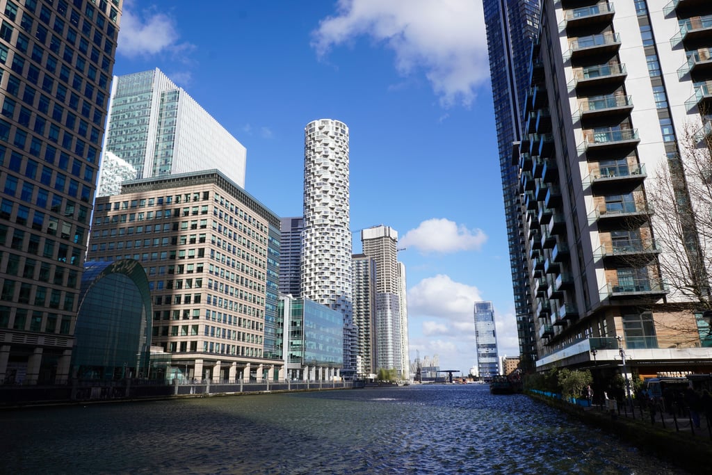 Canary Wharf's modern skyline rises along the waterfront, with glass-covered skyscrapers and a cylindrical tower reflecting against the blue sky.