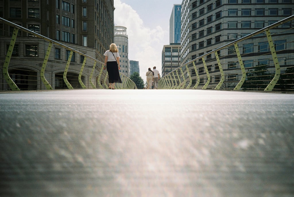 Several people walk across a modern pedestrian bridge with green metal railings, surrounded by tall office buildings and skyscrapers in London's Canary Wharf financial district. The low-angle perspective from the bridge deck emphasizes the urban canyon effect created by the towering glass and concrete structures on either side.