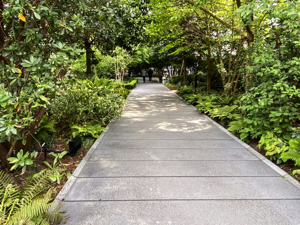 A curved concrete walkway winds through lush tropical vegetation with dense green foliage, ferns, and overhanging tree branches creating a natural canopy overhead. Two people can be seen walking in the distance along the shaded garden path.